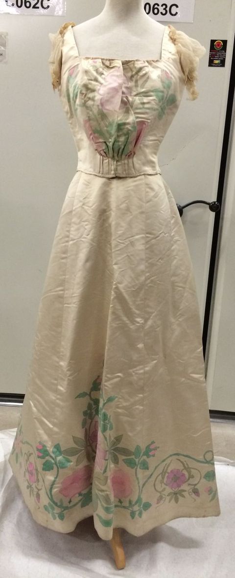Clothing. The short-sleeved bodice of this dress has a square neckline. The skirt falls in an A-line to the floor. The fabric is ivory-white satin so has a sheen. Pink flowers and green stems and leaves are painted on the front of the bodice and around the bottom hem of the skirt.