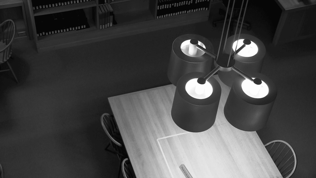 Black and white animation. From overhead, we look down past a chandelier made of four black can lights. The light shines onto a library table below. Photographs of this scene are animated in a short loop so the lights rotate back and forth.