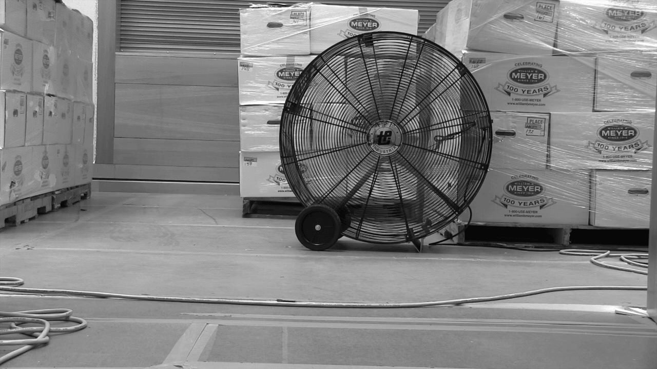 Black and white animation. A black shop fan sits on a floor in front of boxes stacked and wrapped in plastic. Labels on the white boxes include the text, “Meyer” and “100 Years”. Photographs of this scene are animated in a short loop so blades of the fan always spin.