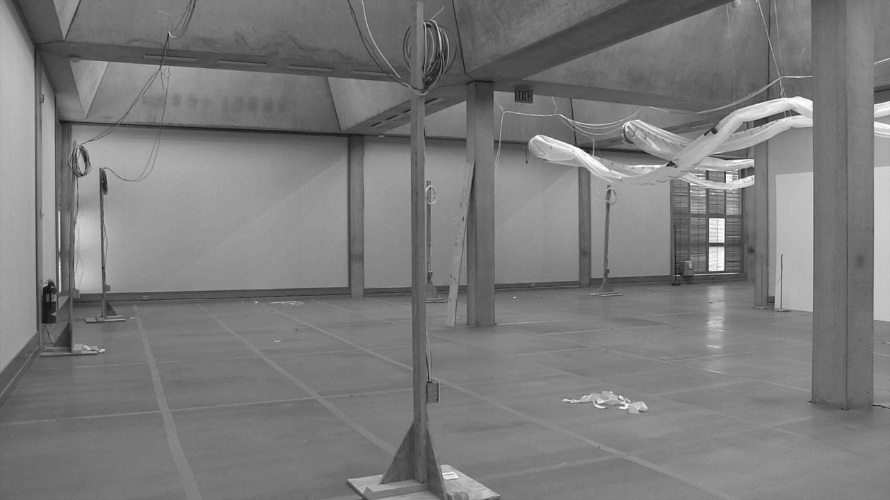 Black and white photograph. Temporary stands hold up wires in a large room. The grid-like ceiling, which opens onto skylights high above, are held up with square columns. Plastic tubing hangs under the ceiling the back right corner of the space.