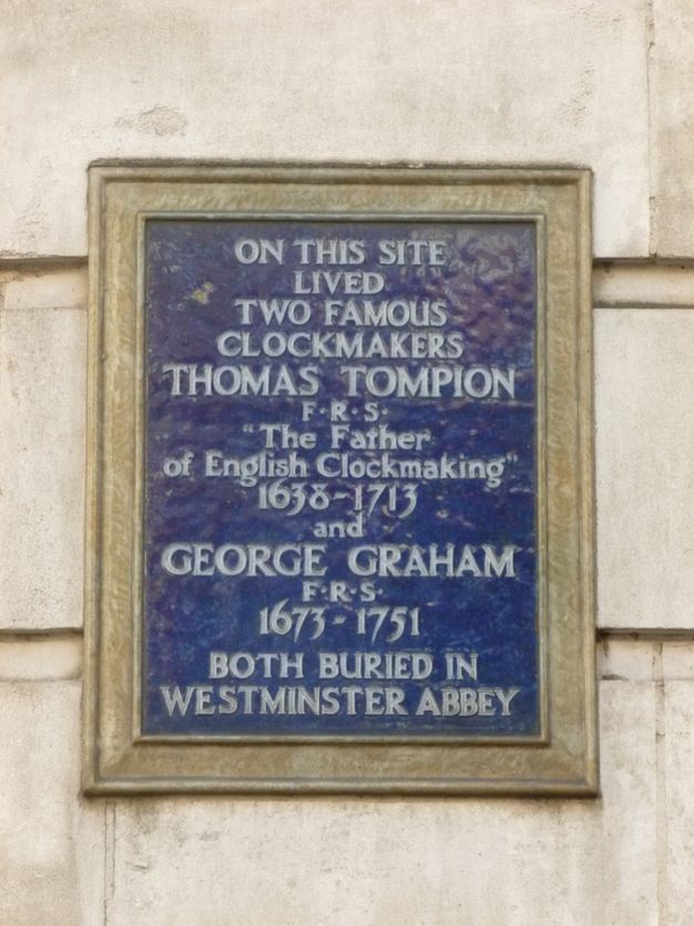 Plaque. A gold-framed sign on the outside of a building reads, “On this site lived two famous clockmakers Thomas Tompion F.R.S. ‘The Father of English Clockmaking’ 1638-1713 and George Graham F.R.S. 1673-1751 both buried in Westminster Abbey”.