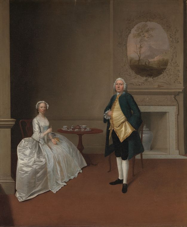 Painting. A woman sits next to a tea set on a round table as a man stands, both looking at us from in front of a hearth in this vertical portrait. Both have pale skin and slightly flushed cheeks. The woman’s wide, satin skirt billows out to her sides, and she wears a bonnet. The man wears a powdered wig and tucks his hand into his gold waistcoat.