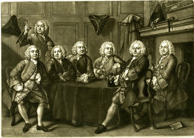 Print. Seven men with almost identical clean-shaven, pudgy, round faces sit or stand around a table in front of a hearth. They all wear white wigs and suits. Tricorn hats hang on the wall behind them or are propped up on the hearth to our right.