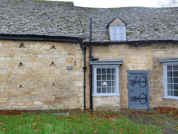 A patch of grassy lawn leads to a brown stone building with a grey slate roof. Under a single, small dormer window, two multi paned windows flank a grey door decorated with ironwork hinges.