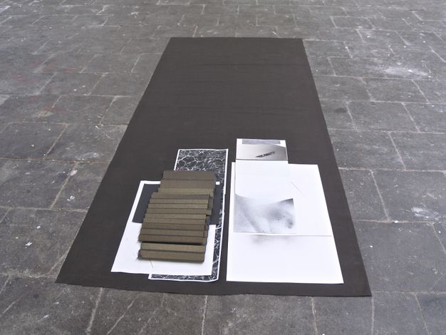 Installation photograph. Several works on paper are layered at one end of a long, rectangular black cloth laid on a grey stone floor. One sheet appears rather large, perhaps A2 size paper. Others are long strips or smaller pieces. Forms and patterns on the papers are in black, white, and shades of grey. Two golden brown pieces are folded like accordions.