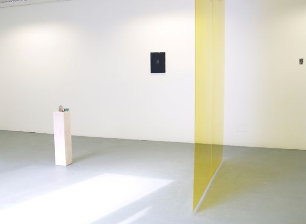 Installation photograph. A translucent yellow panel hovers over a grey floor, hanging perpendicularly to a white gallery wall. Two small works of art hang on the wide expanse of wall beyond the veil. A white, rectangular pedestal holds a few objects, though details are difficult to make out in this photograph.
