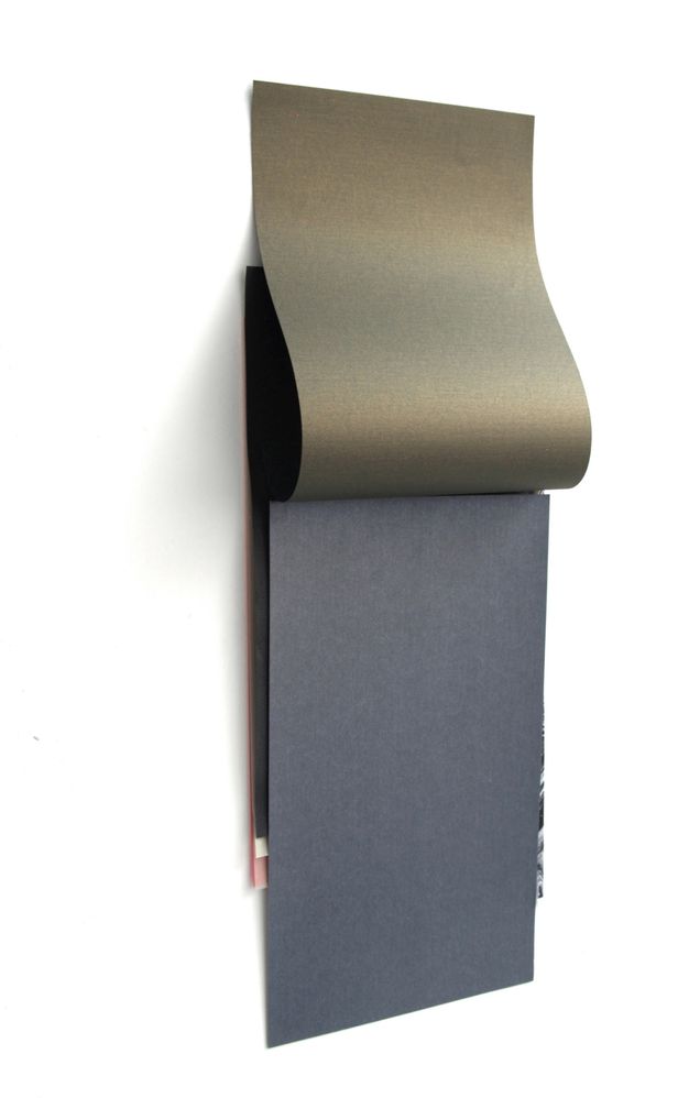 Installation photograph. A charcoal-grey piece of paper hangs on a white wall. A burnished gold-coloured sheet hangs from the wall above the grey, as if the sheets were glued together along the top like a notepad. Slivers of black, white, and pink paper peek out from under the grey sheet.