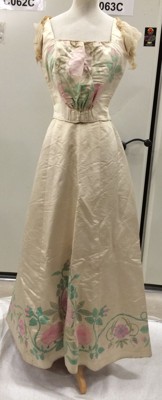 Clothing. The short-sleeved bodice of this dress has a square neckline. The skirt falls in an A-line to the floor. The fabric is ivory-white satin so has a sheen. Pink flowers and green stems and leaves are painted on the front of the bodice and around the bottom hem of the skirt.