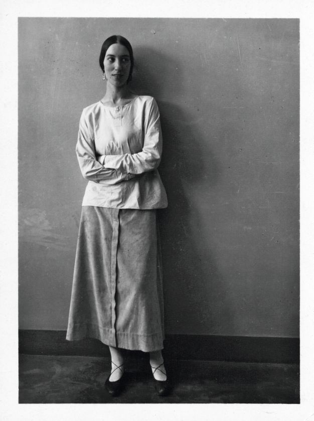 Black and white photograph. A woman stands against a wall with her arms folded across her body, looking off to our right. She has an oval face framed by black hair parted down the middle and pulled back. She wears a loose, long-sleeved shirt and a long skirt.