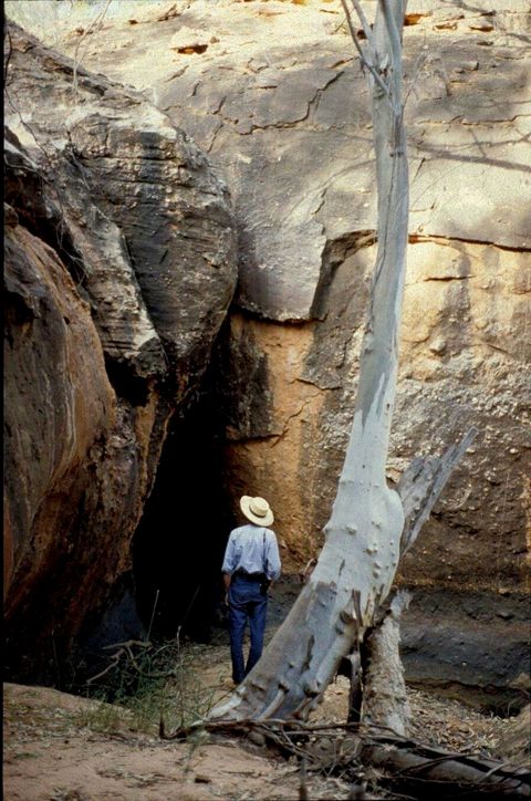 A man wearing a broad-brimmed hat stands facing away from us, looking towards the upside-down, narrow V of a cave entrance. He comes just over halfway up the height of opening. The stone face of the cave fills the picture.