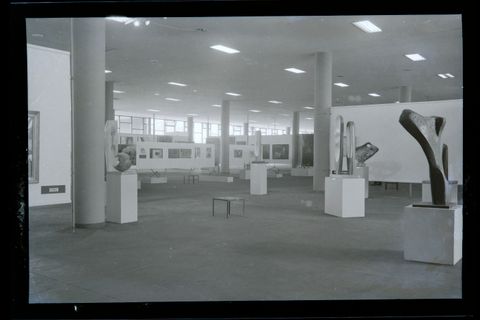 Installation photograph. Black and white photograph shows a window-lined, open space. Abstract sculptures on plinths are arranged throughout the space. In the far corner, works of art are hung on partial white walls.