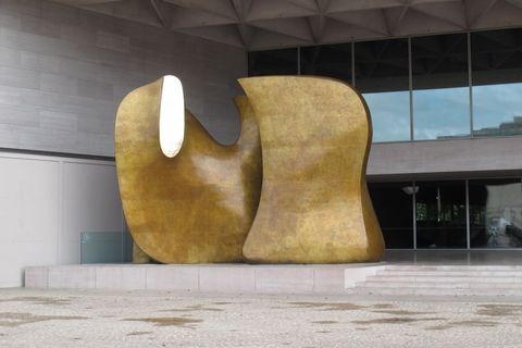 Sculpture. Two rounded bronze forms fill a two-storey, covered entryway. Most of the surface is burnished gold. An oval gleams brightly where the left-hand form seems to have been sliced.