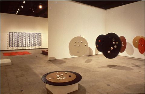 Installation photograph. Six round discs, like tabletops, float in a gallery space, presumably suspended from the ceiling, except for one, which sits on a pedestal. The discs are painted brown, red, or black.