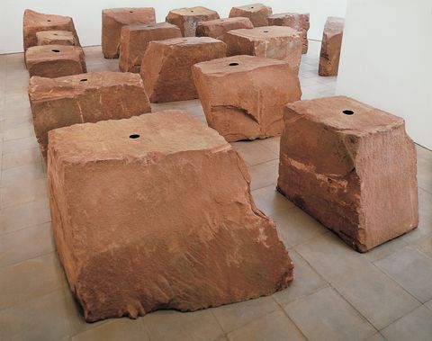 Sculpture. Sixteen roughly cube or rectangular red sandstone blocks sit in rows in a gallery. Each has a black circle at the top centre.