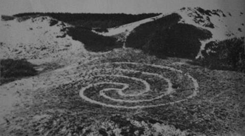 Black and white photograph. A spiral swirls in light lines against a darker, grassy landscape.