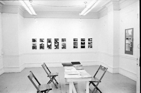 Installation photograph. Black and white photograph of three folding chairs sitting around a long table covered in piles of paper. Photographs and other works of art hang on the gallery walls.