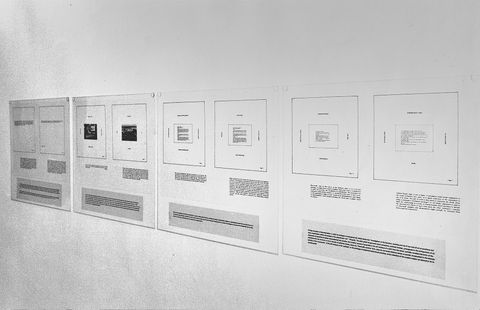Installation photograph. Black and white photograph of four boards hung on a gallery wall in a row. Each board is printed with schematics like floor plans above blocks of text.
