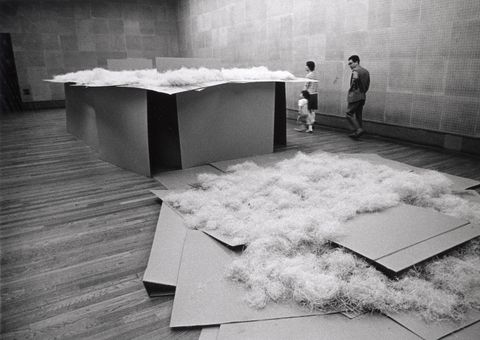 Installation photograph. Black and white photograph shows a table-like sculpture piled with wood shavings. The sides and top are pieces of cardboard stacked like a house of cards. A pile of cardboard and shavings lies on the wood floor, close to us.