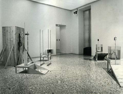 Installation photograph. Black and white photograph of a gallery space with five structures. Each of two fish tanks sit at ramps going down in opposite directions. A cabinet-like structure is across the room.