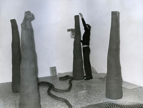 Installation photograph. Four sand-filled, narrow cones stand like sentinels in the corner of a gallery. A man reaches high overhead to drop sand into the cone farthest from us. A thick rope wends across the floor.