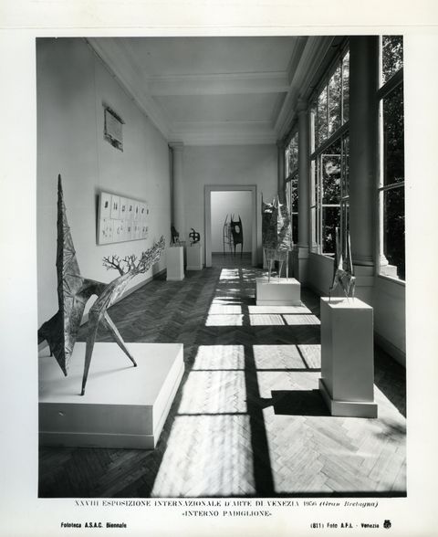 Installation photograph. Black and white photograph of a long, hall-like room lined with windows to our right. Sculptures are set out on plinths along the space, and a multi-piece work is hung on the wall to our left.