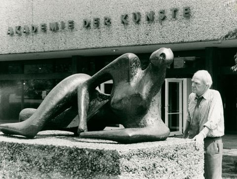 Black and white photograph. A white-haired man with a wrinkled face, Henry Moore, looks under lowered brows at a bronze sculpture of on a waist-high plinth. The sculpture is a stylized reclining female form. The building behind him has the sign, “Akademie der Kunste”.