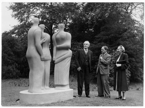 Black and white photograph. Three people stand in a park-like setting next to a light stone sculpture. It has an abstract style and shows three figures with simplified features. The people come up to about elbow height against the sculpted figures.