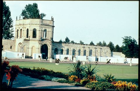 Installation photograph. Abstract and stylized sculptures are displayed on plinths and in an open-air stage set up at the far side of an expanse of grassy lawn. The stage spans the length of a colonnaded stone conservatory.