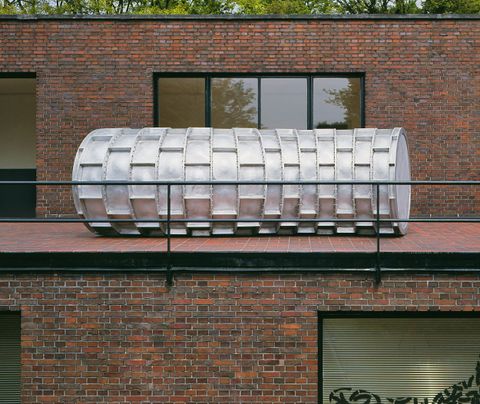 Installation photograph. A long, bright silver pipe made from panels riveted together is on a balcony of a brick building. It spans a double wide window.