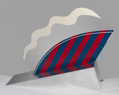 Sculpture. A silver wedge holds up an abstract, fan-shaped form. The bottom half of the fan is striped in navy blue and maroon red, and its curved upper edge is traced by a white neon tube. Above, bands of grey and white ripple like the edge of a pie crust.