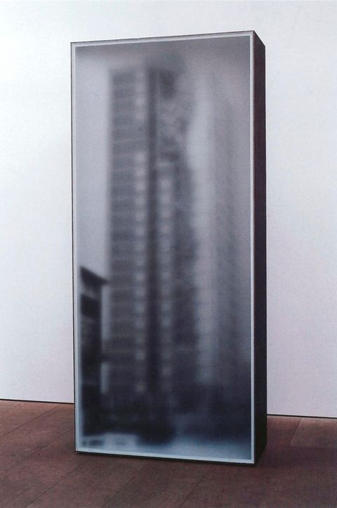 Sculpture. A blurry black and white photograph of a skyscraper is mounted on a tall but shallow, freestanding box in a gallery.