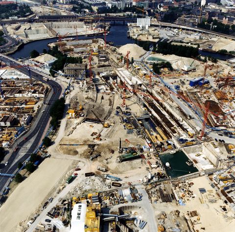 Colour photograph. Aerial view of what appears to be an expansive construction site. A river curves over and through the site, near the top of this photograph. City streets and skyscrapers are visible along the top edge of the image.