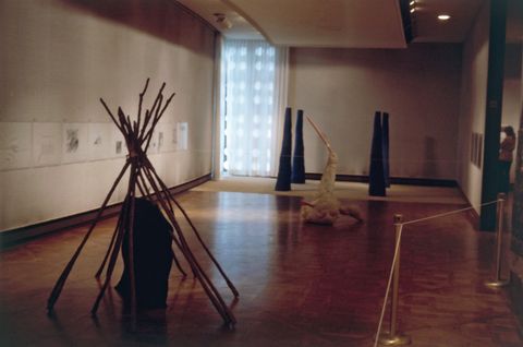 Installation photograph. Sculptural pieces made from wood and other material sit on the floor of a dim gallery. Works of art line the walls.