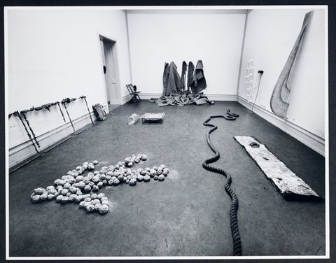 Installation photograph. Black and white photograph of a gallery space. Several abstract sculptures lie on the floor. Another leans against one wall, two are affixed to the opposite wall, and the piece farthest from us appears to be made of cloth hung on the wall and pooling on the floor.