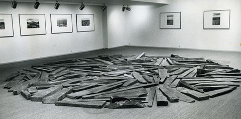 Installation photograph. Black and white photograph of long pieces of slate arranged in a circle. There is no pattern, but the distribution is balanced. Works of art hang on the gallery walls across from us.