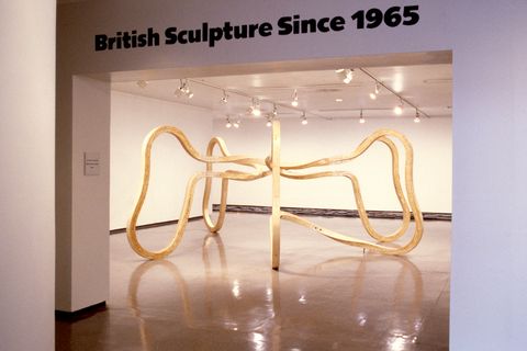 Installation photograph. In the gallery beyond and framed by the opening, an abstract sculpture made from bent wood curves out and down from a central point.