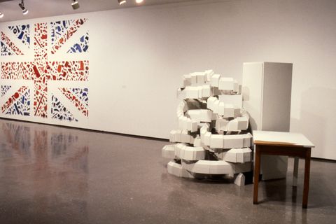 Installation photograph. A Union Jack spans the height of a gallery wall to our left. The red stripes and blue triangles are made from found objects. A work to our right looks like white blocks are stacked in such a way that long rows spiral upwards in a way that defies logic.