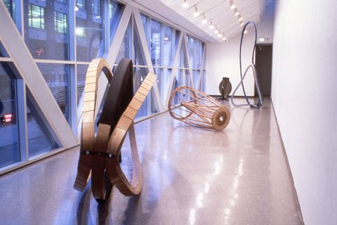Installation photograph. Four abstract sculptures rest on the floor down a long hallway-like space, which is lined with windows to our left. All use curvilinear shapes made of wood or metal.