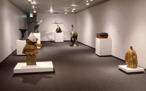 Installation photograph. Seven sculptures are on low or high plinths in a long gallery. Some are abstracted though familiar forms can be found, including a vase, an elephant, and a pile of cloth or clothing.