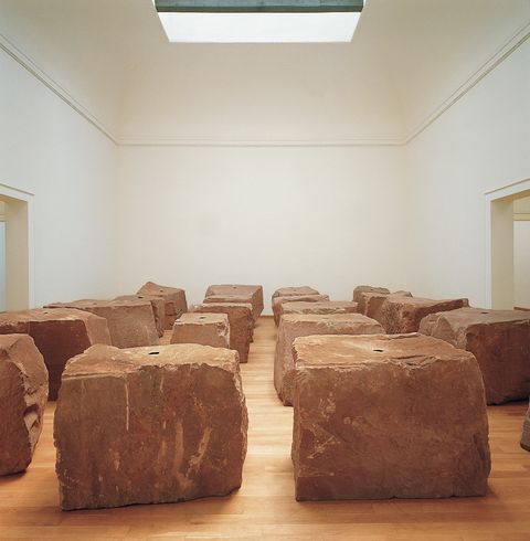 Installation photograph. Twenty roughly cube or rectangular red sandstone cubes sit in rows in a gallery under a skylight. Each has a black circle at the top centre.