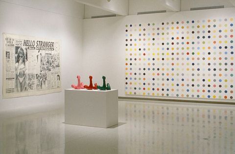 Installation photograph. Two works of art hang on gallery walls and three sculptures share a plinth in the middle of the space. To our left, a dramatically oversized newspaper clipping is titled “Hello Stranger”. A canvas on the wall to our right nearly spans the height of the wall and is painted with a grid of dots, each a single colour.