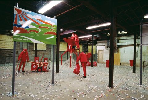 Installation photograph. Five mannequins wearing all red, pick up litter with grabbers, though the whole scene is set in a room with I-beams holding up exposed ceilings. One mannequin dumps a bag into a shredder. A sign near us reads “Scrapheap Services” under a cartoonish landscape.