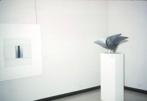A gallery space featuring a minimalist artwork and a sculptural piece. On the left is a framed artwork with a white background and a central vertical element composed of reflective metallic or glass material, creating a sleek and modern aesthetic. On the right is a metallic sculpture displayed on a tall white pedestal. The sculpture consists of curved, intersecting, and pointed forms, showcasing a dynamic and abstract design. The clean, white walls and sparse layout emphasize the contemporary nature of the pieces.