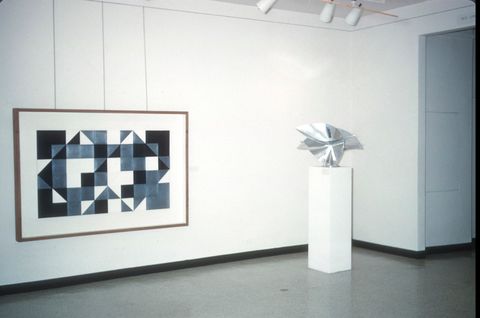 A gallery space with a framed geometric artwork on the left and a metallic sculpture on the right. The artwork features a pattern of interlocking black, white, and gray triangles and squares, creating a bold, abstract design. The sculpture, displayed on a white pedestal, is a shiny, angular form with folded and curved elements, giving it a dynamic, reflective appearance. The background consists of clean, white walls with minimal decor, emphasizing the art pieces.