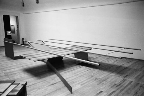Installation photograph. Black and white photograph of an abstract sculpture made of four tubes balanced across four straight and V-shaped panels. The sculpture fills the part of the room we can see.