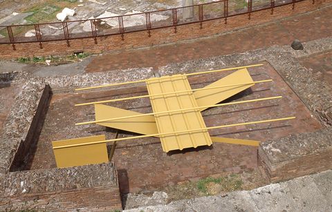 Sculpture. We look down onto a stone courtyard where four metal panels and four tubes, all muted yellow, are balanced to create this abstract sculpture. The four tubes are spaced along a wide strip, which in turn rests on V-shaped strips.