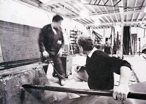 Black and white photograph. Two men work in a studio. One grips a long metal tube and both turn to look at two children standing in the background.