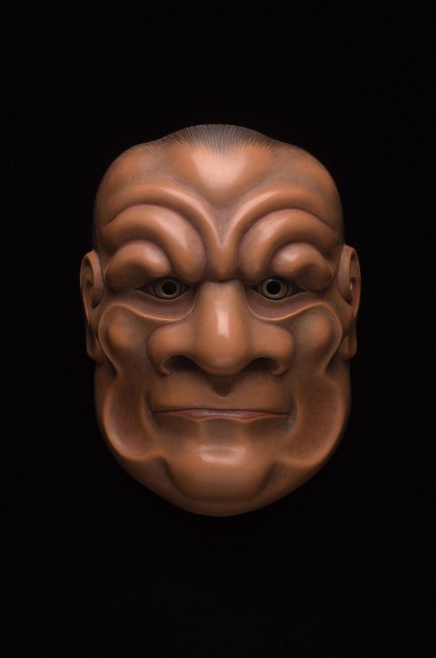 Sculpture. Wooden mask of a stylized man’s face. Wrinkles curving from the wide nose up over eyes and those lining the cheeks are exaggerated. The upper lip disappears over a thin lower lip. The skin is light brown.