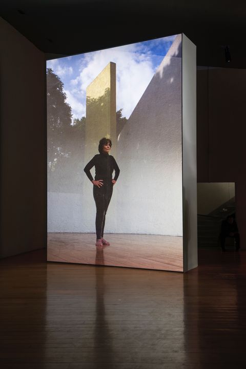 Installation photograph. A film is projected onto a floating wall. This still shows a woman wearing all black standing with her bare feet together and her hands on her hips. Projected behind her is a view of outdoor architecture or a sculpture.