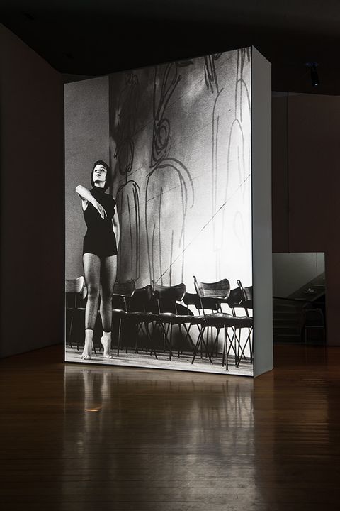 Installation photograph. A black and white film is projected onto a floating wall. The still shows a woman with dark hair wearing a black leotard and standing on tiptoes next to rows of wooden chairs. One elbow is gracefully lifted, mid-dance.
